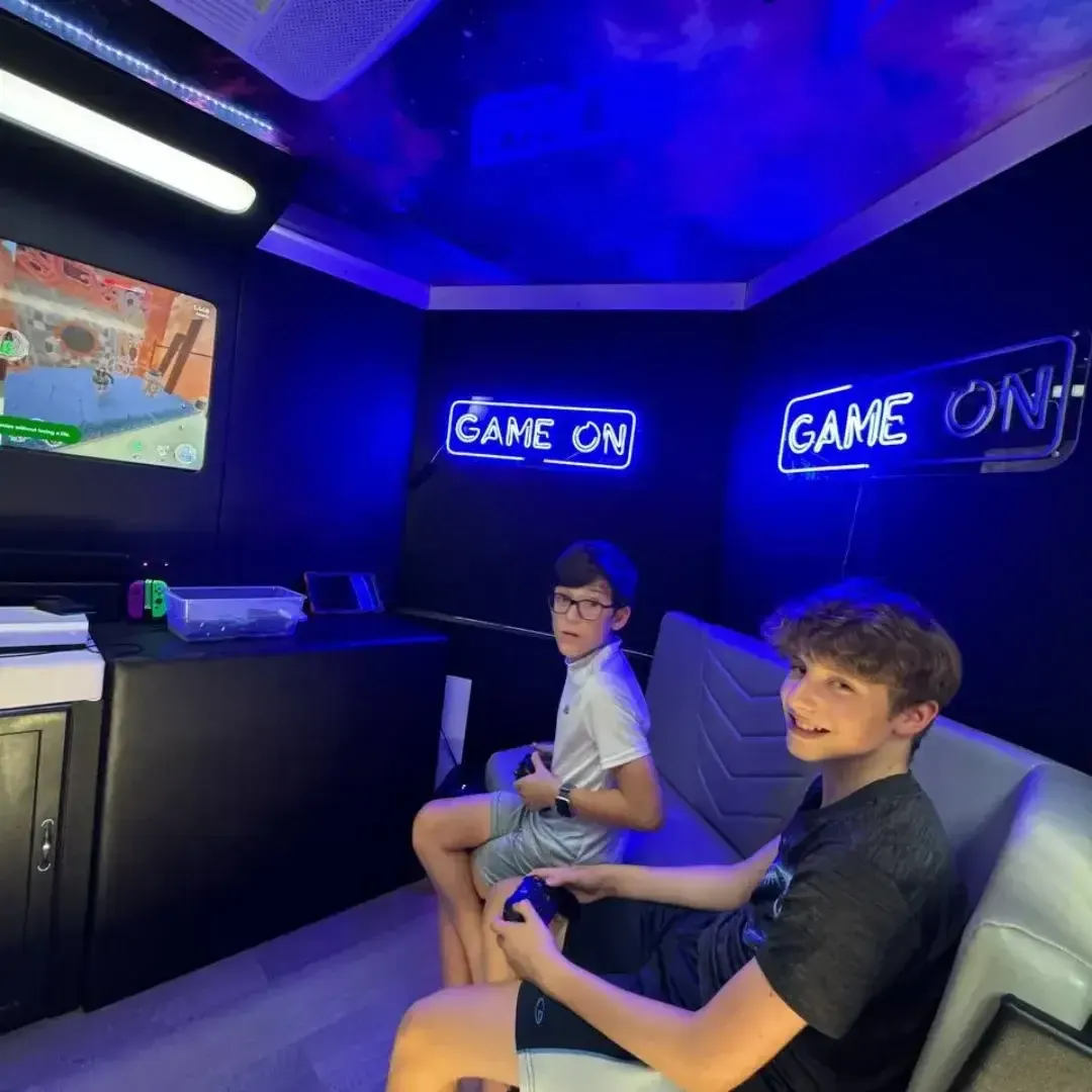 Two kids playing console games under a blue GAME ON neon sign in the gaming truck